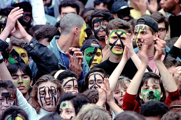 Foto colorida de pessoas com rosto pintado nas cores verde, amarelo e preto. Algumas erguem as mãos e batem palmas.